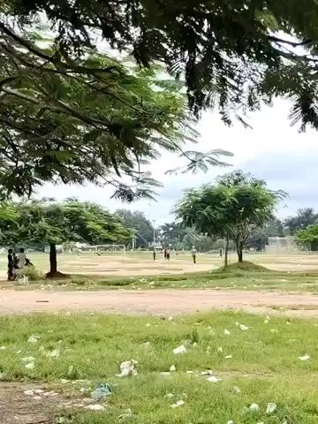 The video shows a grassy field with scattered litter, likely plastic bottles and wrappers, on the ground. In the background, there are trees, some buildings, and a cloudy sky. There are a few people in the distance, appearing to be walking or playing. One person is seen on a dirt path in the middle ground. The scene appears to be a park or an open recreational area.