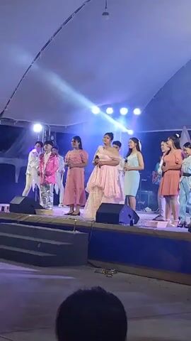 A group of people, including at least five young women and two young men, are standing on a stage under bright lights. One young woman in a pink dress is holding a microphone and appears to be singing. She is surrounded by others in similar festive attire, including one young man in a sparkly pink jacket and silver pants. The stage is set up outdoors, with a large white tent overhead. The lighting is dramatic, with spotlights creating a hazy, atmospheric effect. The overall impression is of a performance or celebration taking place at night.