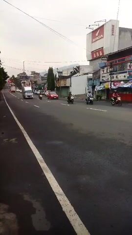 The video shows a street scene. A two-lane asphalt road runs diagonally from the bottom left to the upper right of the frame. White lane markings are visible on the road. The right side of the road is lined with buildings. One building has a large red and white sign that says "HONDA" at the top. Other buildings appear to be shops or businesses. There are various vehicles on the road, including cars, trucks, and motorcycles. Several motorcycles are in motion, with riders on them. A white truck is moving away from the camera in the left lane. A red car is also visible on the road. The sky is overcast. Trees and utility poles are present along the side of the road. The overall impression is of a moderately busy street in a town or city.