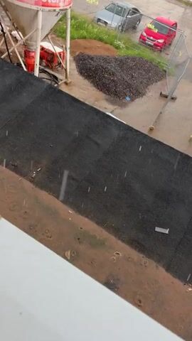 The video shows a construction site with rain falling. A large cement silo is tilted precariously on its stand. Various construction materials and equipment are visible, including a pile of gravel and what appears to be part of a temporary structure or barrier. In the background, there is a street with several cars parked and driving in the rain. A multi-story building can be seen to the right of the frame. The rain creates ripples in puddles on the ground and streaks across the camera lens.