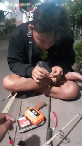 A person is sitting on a wooden surface, hunched over and focusing on their hands. They appear to be tying a fishing line, as evidenced by the fishing gear in the foreground, including a fishing rod, a tackle box, and a bobber. The person is wearing a black long-sleeved shirt and dark shorts, with their legs crossed. The setting seems to be outdoors, possibly near a body of water, given the presence of foliage and the faint reflection in the background. The lighting suggests it is evening or nighttime, with some artificial lights visible in the distance. Another person's hand is visible in the lower left corner of the frame, also holding a fishing line.