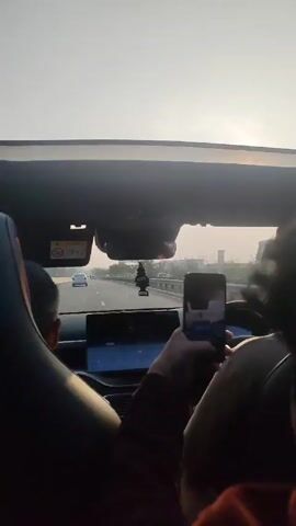 The video shows a view from inside a moving car on a highway. The passenger side of the car is primarily visible. The person sitting in the passenger seat is using their mobile phone to film. Outside, other cars are visible on the road. The sky is bright and hazy.
