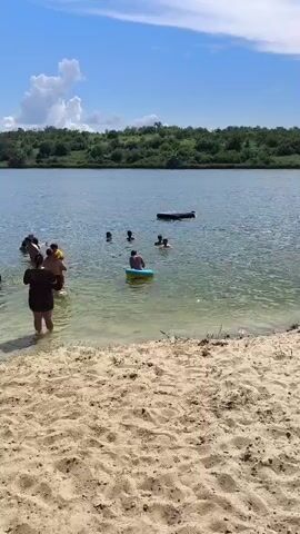 A group of people are in a lake on a sunny day. Some are in the shallow water near the sandy shore, while others are swimming further out. One person is on a wooden dock and appears to be preparing to jump into the water. Another person is holding a yellow inflatable ring in the water. In the background, there is a line of green trees and a blue sky with white clouds. The water is clear and calm. The scene suggests a casual summer outing with friends and family enjoying the water.
