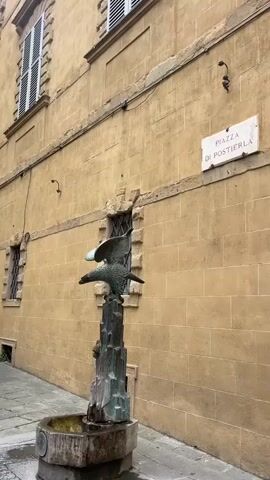 The video shows a bronze statue of an eagle with outstretched wings perched atop a stone pedestal with a small basin at its base, forming a fountain. The fountain stands in front of a tan-colored brick building with a street sign reading "Piazza di Postierla" attached to the wall. The building has windows with white shutters and decorative details around the openings. The street is paved with stone tiles, and the scene is lit by natural daylight. The camera angle is from a low perspective, looking slightly upward at the statue and building. There are no people visible in the frame. The water is trickling from the statue to the fountain.