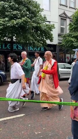 A group of people are walking in a parade on a street. Many of them are wearing traditional Indian clothing, such as saris and kurtas. Some people are wearing garlands of flowers around their necks. There is a car parked on the side of the street, and buildings line the street in the background. The people appear to be singing and dancing.
