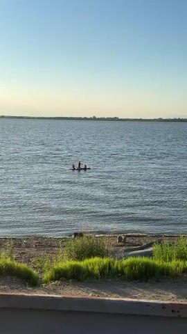 A wide shot captures a calm body of water under a clear blue sky. In the middle of the water, a small red raft or kayak carries three individuals. One person stands and appears to be paddling, while the other two are seated. The water is slightly rippled. The shoreline in the foreground is composed of sand and pebbles, with patches of green grass and some vegetation. In the distance, the horizon is marked by a faint line of land or trees. The lighting suggests it is either early morning or late afternoon.