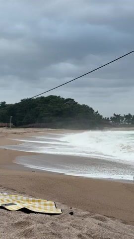 The video shows a cloudy beach scene. The sand is light brown, and there are footprints in the foreground. A yellow and white checkered blanket is spread on the sand. The ocean waves are white and foamy, crashing onto the shore. In the background, there is a hill covered in green trees, with some structures visible on top. A dark line, possibly a cable, stretches diagonally across the top of the frame.
