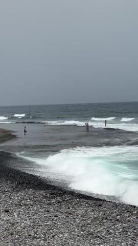 The video captures a scenic view of a rocky beach with the ocean in the background. The beach is composed of dark, smooth pebbles. Several people are visible in the shallow waters and on the rocky outcrops, enjoying the day. In the distance, gentle waves are breaking, and a few individuals appear to be surfing or preparing to surf. The sky is overcast, suggesting a cloudy or hazy day, possibly in the morning or late afternoon. The water is a clear, turquoise hue where the waves are breaking, contrasting with the darker, deeper ocean. The overall mood is one of leisure and enjoyment of the coastal environment. There is no visible text in the media file. The location context suggests this scene is in Arona, Spain, which is known for its diverse coastlines.