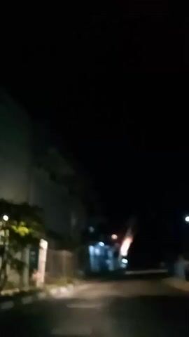 The video appears to be recorded from inside a moving vehicle at night. The camera is pointed towards the right side of the road, where a building with lights is visible. There are several flags attached to the building, illuminated by lights, and they are waving. The road is dark and appears to be asphalt. Trees and a fence are also visible along the side of the road. The sky is completely dark.