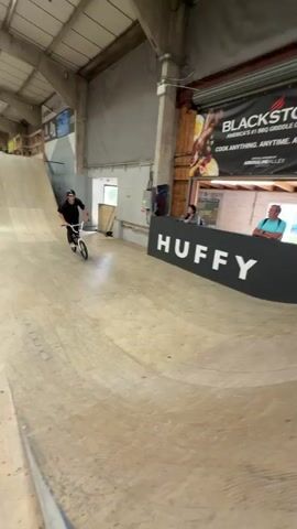 Here is a description of the video:

In an indoor skateboarding park, a young man performs a bike trick. He rides a silver BMX bike through a wooden ramp. He wears a black shirt and helmet. He performs a jump, becoming airborne, and then lands smoothly. The park features a high ceiling with skylights. There's also a banner and other park infrastructure visible. Another person is seen standing behind a counter in the background.