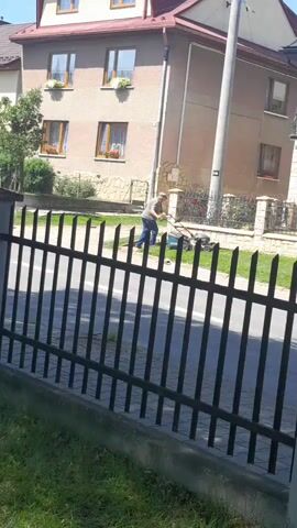A person is mowing a lawn with a push mower in front of a multi-story building. The person is wearing a light-colored shirt and dark pants. The lawn is bordered by a black metal fence. The building appears to be a residential house. The day is sunny.