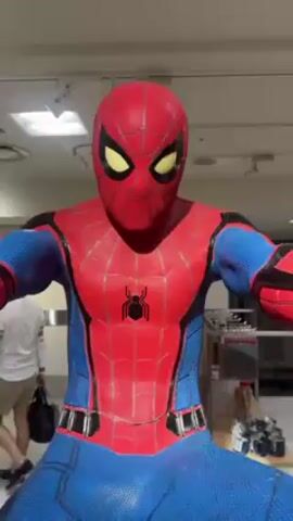 The video shows a full-body statue of Spider-Man in a crouching pose. The statue is detailed with red and blue coloring and is positioned in what appears to be a retail environment. Shoppers browse in the background. The camera slowly zooms out, offering a full view of the statue and its surroundings. The statue is positioned on a black base.