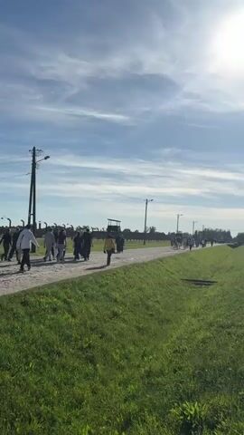 The video shows a sunny outdoor scene with a grassy area in the foreground. A gray gravel path runs parallel to a set of train tracks. A group of people is walking along the path. The sky is blue with some wispy clouds and a bright sun. In the distance are some utility poles and possibly structures, but details are unclear.