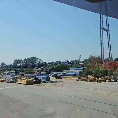 The video shows a large outdoor storage area. In the background, numerous cars are parked on tiered levels of what appears to be a junkyard or salvage yard, with many of them being SUVs and trucks. In the foreground, there are stacks of construction materials, including lumber, metal pipes, and other building supplies, arranged in neat piles. The sky is clear and blue, suggesting a sunny day. The ground appears to be paved or gravelly. The overall scene depicts an organized but extensive storage facility for both vehicles and building materials.