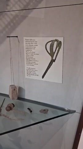 The video displays a museum exhibit. In the foreground, a glass shelf holds various small, rough-hewn objects, possibly artifacts. Among these objects are pieces of twine, a small wooden spool, and what appears to be a small, dark, cylindrical object. Behind the shelf, on the wall, is a framed text and an image of a stylized bronze tool, resembling scissors or shears. The text, written in Italian, discusses early agricultural tools and women's crafts, referencing a bronze fibula dated to the 8th century BC. To the left of the framed exhibit, a thin string hangs from the ceiling, suspending a small, weathered stone object. The overall lighting suggests an indoor, possibly dimly lit museum environment, with shadows cast on the wall from the exhibit elements.