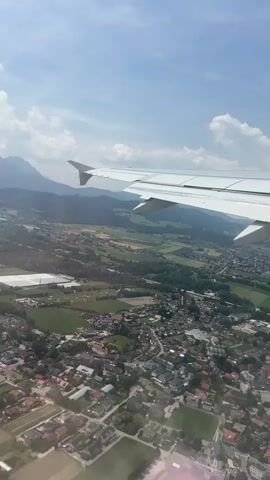 This aerial footage captures a view from an airplane window, showcasing a scenic landscape on a sunny day. The primary subject is the airplane's wing, a prominent white object extending into the frame from the right, with its distinctive wingtip and visible flap mechanisms. Below the wing, a sprawling town is visible, characterized by numerous houses with red-tiled roofs, interspersed with green spaces, fields, and some industrial or commercial buildings.

The scene unfolds over a valley, with lush green fields and darker patches of trees forming a rural-agricultural backdrop. In the distance, imposing mountains rise, their peaks partially obscured by a light haze, suggesting a vast and mountainous region. The sky above is a vibrant blue, dotted with fluffy white clouds, indicating fair weather. The sunlight casts bright illumination, creating a cheerful and clear atmosphere.

The activity taking place is likely a plane in ascent or descent, given the low altitude over the town and the clear visibility of the landscape. The perspective suggests the plane is moving, with the ground appearing to glide by. The time of day appears to be daytime, likely late morning or early afternoon, due to the strong sunlight and the bright sky.

No discernible text is visible in the footage. The overall emotion conveyed is one of travel and the beauty of natural and human-made landscapes viewed from above, evoking a sense of journey and discovery. The specific location context of Siezenheim, Austria, further enhances this understanding, suggesting a view over a European town nestled within a mountainous region.