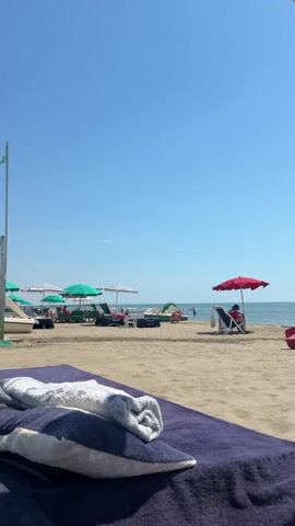 The video shows a beach scene on a sunny day. In the foreground, a lounge chair is covered with a dark blue towel and two pillows, one blue and one white, with a folded gray towel on top of them. The sand extends into the midground where several beach umbrellas, some green and some white, are set up. A few lounge chairs are visible under these umbrellas. To the right of the frame, a single red umbrella covers a lounge chair with a person sitting in it, facing the water. Beyond the beach, the ocean stretches to the horizon under a clear blue sky. There are a few small boats or watercraft visible in the distance on the water. The overall impression is one of a tranquil beach day.