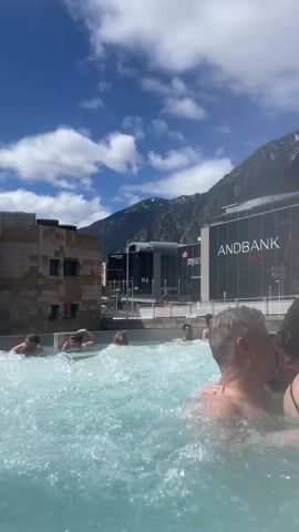 A brightly lit daytime outdoor shot shows multiple people of varying ages enjoying a large pool with strong bubbles.  The pool is surrounded by building structures on one side and mountains on the other.  The sky is blue with fair-weather cumulus clouds.  Some people are clearly visible, while others are partially obscured by the bubbles and splashing water.  A couple in the foreground interact playfully with each other.
