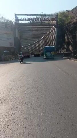 A perspective shot from a moving vehicle shows a road leading under a large overpass. The overpass has "SELAMAT DATANG DI KABUPATEN BANDUNG" written on it. Trucks and motorcycles are traveling on the road. The overpass has a curved, ribbed structure supporting it. The road surface is asphalt and appears worn in places. The sky is visible above the overpass, and it is bright, suggesting it is daytime. The surrounding environment appears to be a mix of rock formations and foliage on the right side of the road.