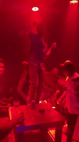 A group of people are gathered in a dimly lit room with red lighting. In the center of the room, a man is standing on a table, dancing. He is wearing a dark shirt and light-colored pants. There are drinks on the table. Other people are standing around the table, some watching the man, others talking. The atmosphere appears to be that of a club or party.