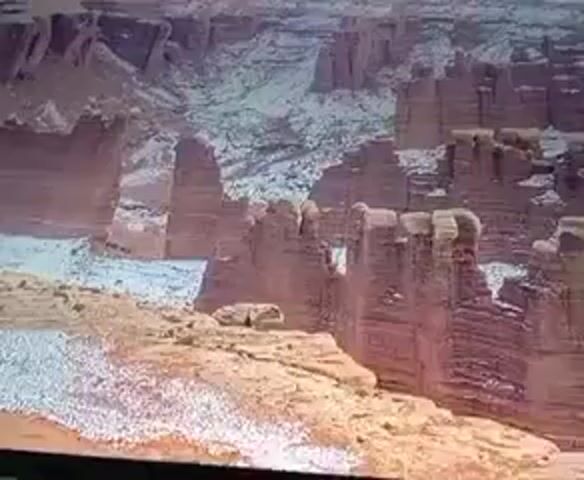The video shows a wide shot of a desert landscape with rock formations. The rock formations are tall and columnar, with layered sedimentary rock visible. There is some snow on the ground and on the rocks, suggesting a cold environment. The colors of the rocks are primarily reddish-brown and tan, with white areas where snow is present. The sky is not visible in the frame. The overall impression is of a rugged and natural landscape.