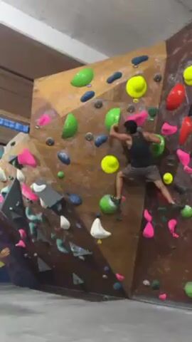 A man is bouldering on an indoor climbing wall. The climbing wall is multi-faceted with different colored holds. The man wears a tank top and shorts. He has his back to the camera as he reaches upwards on the wall. Another man walks into the frame in the lower-left, as does another man in red shorts. The climbing gym has a high ceiling and concrete floor.