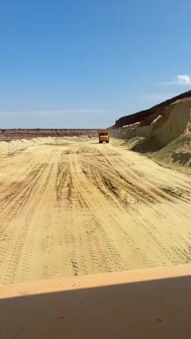 A wide shot captures a construction site under a clear blue sky. The terrain is sandy and uneven, with tire tracks clearly imprinted on the ground, suggesting heavy machinery activity. On the right side of the frame, a dirt embankment rises steeply, showcasing layers of sand and reddish-brown soil. In the distance, a large yellow dump truck is seen driving away from the camera along the dusty road. The truck's bed is raised, indicating it might be carrying a load. Other construction vehicles appear to be present further back in the scene. The camera appears to be positioned within another vehicle, with a portion of its orange-colored interior visible at the bottom of the frame. The overall impression is one of ongoing large-scale earthmoving operations.