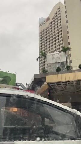The video shows a view from inside a car, looking out through the rain-streaked windshield. In the background, a tall, beige building with "ICBC" written near the top is visible, along with some palm trees. To the left of the building, a large green billboard advertising "Fusion Cuisine & Lounge" is partially visible. The sky is overcast and appears to be raining.
