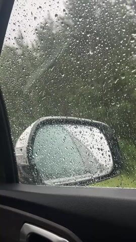 The video shows the interior of a car on a rainy day. Raindrops are visible on the car window, blurring the view of the outside world. A side mirror is also visible, covered in raindrops. The background is a blurry green landscape, suggesting trees or foliage. The interior of the car is dark, with a portion of the car door visible in the foreground. The overall atmosphere is moody and tranquil, typical of a rainy drive.