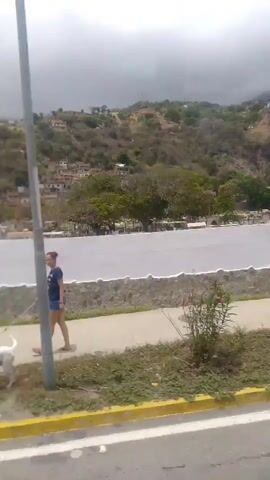 The video shows a moving view of a roadside scene under an overcast sky. The foreground includes a yellow-painted road line, vegetation, and light posts. Beyond this, a white wall with a light green arched entrance suggests a cemetery or similar structure. In the background, a hillside is visible with buildings scattered across it. As the camera moves, buildings and people come into view. Several people are walking on a sidewalk near a building with a blue window frame. The overall impression is a shot taken from a moving vehicle driving past a roadside scene in a somewhat hilly, populated area.