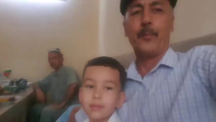 The video displays a slightly blurry, close-up, first-person perspective of a man and a young boy. The man, who is in the foreground, is wearing a striped collared shirt and a dark cap, and has a mustache. He is holding a young boy in front of him, who is wearing a white collared shirt. The boy is looking directly at the camera with a neutral expression. Behind them, an older man is seated at a table. The table is laden with various food items, including fruit and what appears to be bread, as well as teacups. The background consists of a light-colored wall and a window with blinds. The overall scene suggests a casual gathering or meal.