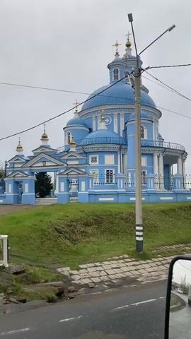 The camera is positioned inside a moving vehicle and is facing towards a blue and white church. The church is situated on a raised grassy area beside the road, supported by a low stone retaining wall. The church has a central dome with multiple smaller domes and towers. The sky is overcast and grey. A utility pole is visible in the foreground. The video is short and appears to be a quick recording from inside a car as it passes by the church.