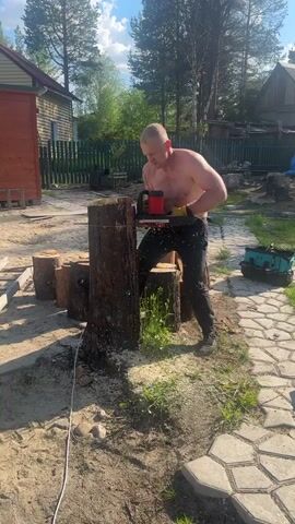 A shirtless man in black pants and yellow gloves uses a red and black chainsaw to cut wood. He stands in front of a tree stump, with sawdust flying around him. Other cut wood logs are on the ground to his left. A house with a red facade is visible in the background, along with a fenced area and trees. A paved path with hexagon-shaped stones is on the right side of the frame. The sky is blue with some clouds.