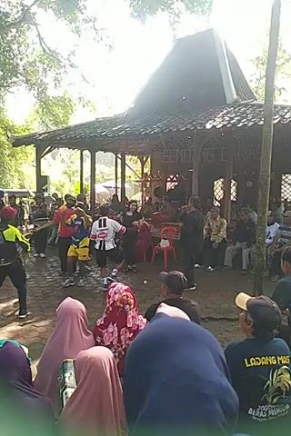 Here is a brief description of the video:

The video shows a scene under a wooden pavilion with a tiled roof. A man in a black suit is standing and speaking into a microphone, likely performing or giving a speech. A band is set up behind him. Several people are gathered around the pavilion, some sitting, some standing, and some looking towards the man speaking. The setting appears to be outdoors with bright sunlight.