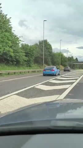 The video shows a view from inside a vehicle driving on a highway. The highway has multiple lanes marked with white lines and arrows. Lush green trees and foliage line the sides of the road, with some overpasses and streetlights visible. In the foreground, the dashboard and windshield of the camera vehicle are partially visible. A blue BMW is driving in the adjacent lane, followed by a dark-colored car. The camera vehicle passes under an overpass and continues on the highway. Further ahead, a white van and other vehicles are seen on the road. The sky is overcast with clouds.