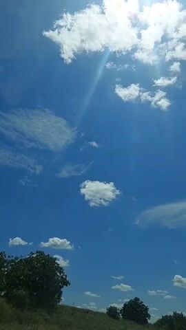 This is a vertical video shot from a low angle looking up at a bright blue sky. There are scattered white clouds, with some appearing wispy and others more puffy. Sunlight is streaming down from the top right of the frame, creating visible sunbeams and highlighting some of the clouds. At the bottom of the frame, there is a grassy slope with several trees and bushes. The overall impression is a sunny day in an outdoor, natural setting.