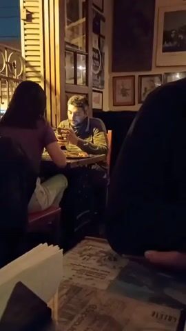 The video shows a dimly lit restaurant or bar, filled with people. The camera focuses on a man with dark hair and a mustache sitting at a table with a menu, while other patrons can be seen in the background. The walls are adorned with a multitude of framed pictures and posters. There are some tables and windows visible, and the atmosphere appears bustling, with people talking and moving around.