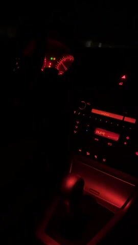 The video shows the interior of a car at night, illuminated by red ambient lighting. The dashboard displays are visible, including a speedometer and a radio with a temperature reading of 16.0°C. The gear shifter is also visible, glowing red. The overall scene is dark, with the red lights creating a moody atmosphere. There are no people or interactions visible in the video.
