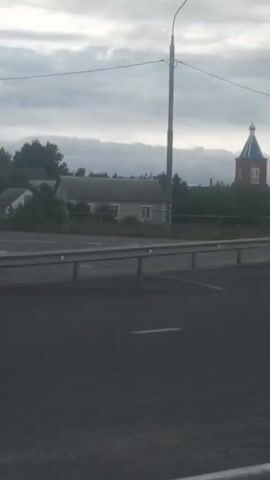A car is driving on a road, with a church visible in the distance. The sky is cloudy. The car is moving forward and then the scene cuts to a different angle, showing the car continuing to drive down the road.