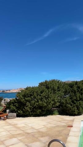 The video shows a picturesque outdoor scene on a bright, sunny day. The camera begins facing a paved patio area with a green bush on the right, a green lounge chair on the left, and a glimpse of turquoise water and rocky islands in the background. The sky is a deep, cloudless blue. As the camera pans slightly to the left, a white gazebo-like structure comes into view, providing a partial frame to the scene. The focus then shifts more towards the water, revealing a clearer view of the rocky coastline, the turquoise sea, and a distant mountain range with snow-capped peaks. The lounge chair remains visible on the patio. There are no people visible in the video. The overall impression is that of a serene, coastal vacation spot.