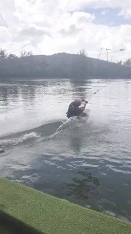 A person is wakeboarding on a lake. They are being pulled by a cable system, which is visible overhead. The wakeboarder is in the process of performing a trick, with spray coming off the water around them. In the background, there are trees and some structures, possibly part of a wake park. The sky is cloudy, and the overall lighting suggests it is daytime. The scene appears to be set in a recreational area, likely Tambon Mai Khao, Thailand, given the provided location context. There is no visible text in the media.
