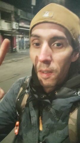 A man wearing a dark blue jacket, a black hoodie, and a light brown beanie is shown in a close-up selfie-style video. He is outdoors at night, with a street and buildings visible in the blurred background. He appears to be speaking and gesturing with his right hand, with his index finger extended. A red object is clipped to his jacket. The video is filmed from a low angle, looking up at the man.