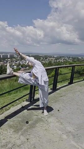 A woman in athletic attire is performing a series of stretches and poses on a paved area next to a railing. She is facing away from the camera for most of the video, with her back and right side visible. Her body is positioned to the left of the frame, with her legs extended and her torso bent forward. In the background, a city or town is visible, nestled in a valley surrounded by rolling hills. The sky is blue with scattered white clouds. The woman is barefoot or wearing light-colored shoes. Her movements are fluid and controlled as she transitions between different poses, demonstrating flexibility and balance. She appears to be in a park or elevated viewpoint, given the railing and the expansive view of the landscape.