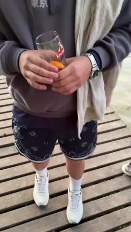 The video shows two people walking towards the end of a wooden pier on a lake. The person in the foreground is holding a glass of amber liquid in their right hand and a white towel over their left shoulder. They are wearing a gray hoodie, navy shorts with a white pattern, white socks, and white sneakers. The other person is walking behind them to their right and is wearing a brown jacket, black leggings, and gray sneakers, and is holding a pink cell phone. The pier extends into the lake, and there are reeds growing along the water's edge. The camera pans upwards to show a sunset over the lake. The sky is a mix of orange, pink, and gray clouds.