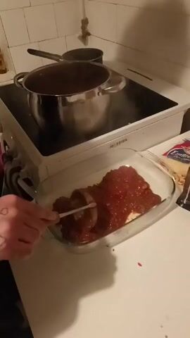A person in a white and black patterned sweater is using a metal spoon to evenly spread a dark red sauce over a white base in a rectangular glass baking dish. The dish sits on a white countertop next to a package of shredded cheese. Behind the dish is a white stovetop with a large stainless steel pot and a smaller saucepan on it. Steam rises from the large pot. The kitchen has white tiled walls.