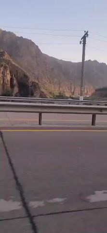 A white sedan is visible from its side as it travels on a paved road. The car has tinted windows and a silver rim on the rear wheel. In the background, a steep, rocky mountain slope is illuminated by sunlight. A metal guardrail separates the road from the mountain. A utility pole stands on the side of the road near the mountain. The sky is clear and blue.