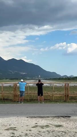 Two men stand facing a runway, watching an airplane take off. The man on the left wears a blue shirt and shorts and a white cap. The man on the right wears a black shirt and shorts. They are both standing behind a fence. The runway is visible in the background, with mountains behind it. The sky is partly cloudy. The airplane is in the air, ascending.