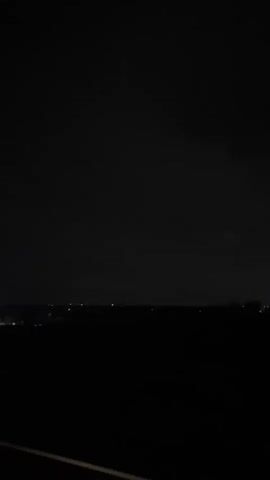 The video shows a dark, nighttime scene with a faint horizon line dotted with distant lights. The foreground is black, indicating darkness or perhaps a shadowed surface like a road. The sky is also dark, with subtle variations in shade suggesting clouds or atmospheric conditions. There are no people or clearly defined objects visible in the frame, and no discernable actions or interactions are taking place. The overall impression is one of a vast, dark expanse, possibly a landscape viewed from an elevated position or during a drive.