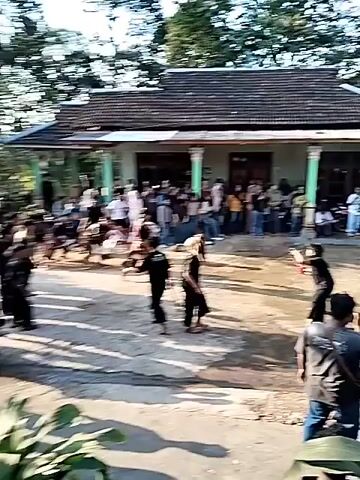 The video captures a vibrant outdoor cultural performance in what appears to be a village courtyard. A group of performers, mostly dressed in black, are engaged in a dynamic dance or martial arts demonstration. Some are wielding whips, which they crack energetically, while others move with impressive agility, performing kicks and jumps. The central focus of the performance seems to be a large, intricately decorated effigy, possibly a dragon or a mythical creature, being carried and manipulated by several individuals. A crowd of onlookers, comprising men, women, and children, are gathered on the porch and in the yard of a traditional-looking house, observing the spectacle with evident interest. The setting is rustic, with dirt ground, a modest building with a tiled roof, and lush green trees in the background, suggesting a rural or semi-rural environment. The lighting indicates it is daytime, likely late afternoon, with the sun casting long shadows.