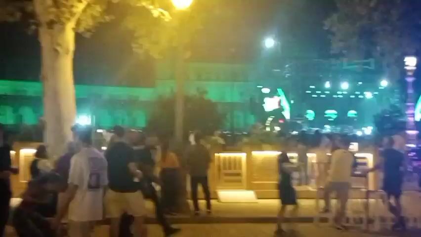 The video shows an outdoor scene at night. In the background, there is a large building lit with green lights, suggesting an event or concert is taking place. In front of the building, there is a stage with bright lights and screens displaying imagery. Several people are gathered in the foreground, some standing and talking, others appearing to be waiting in line. There are tables and chairs set up, possibly for a bar or seating area. The overall atmosphere appears festive and social, with ambient lighting contributing to the nighttime ambiance. Trees are visible, with some illuminated by artificial lights. The video is shaky and appears to be handheld footage.
